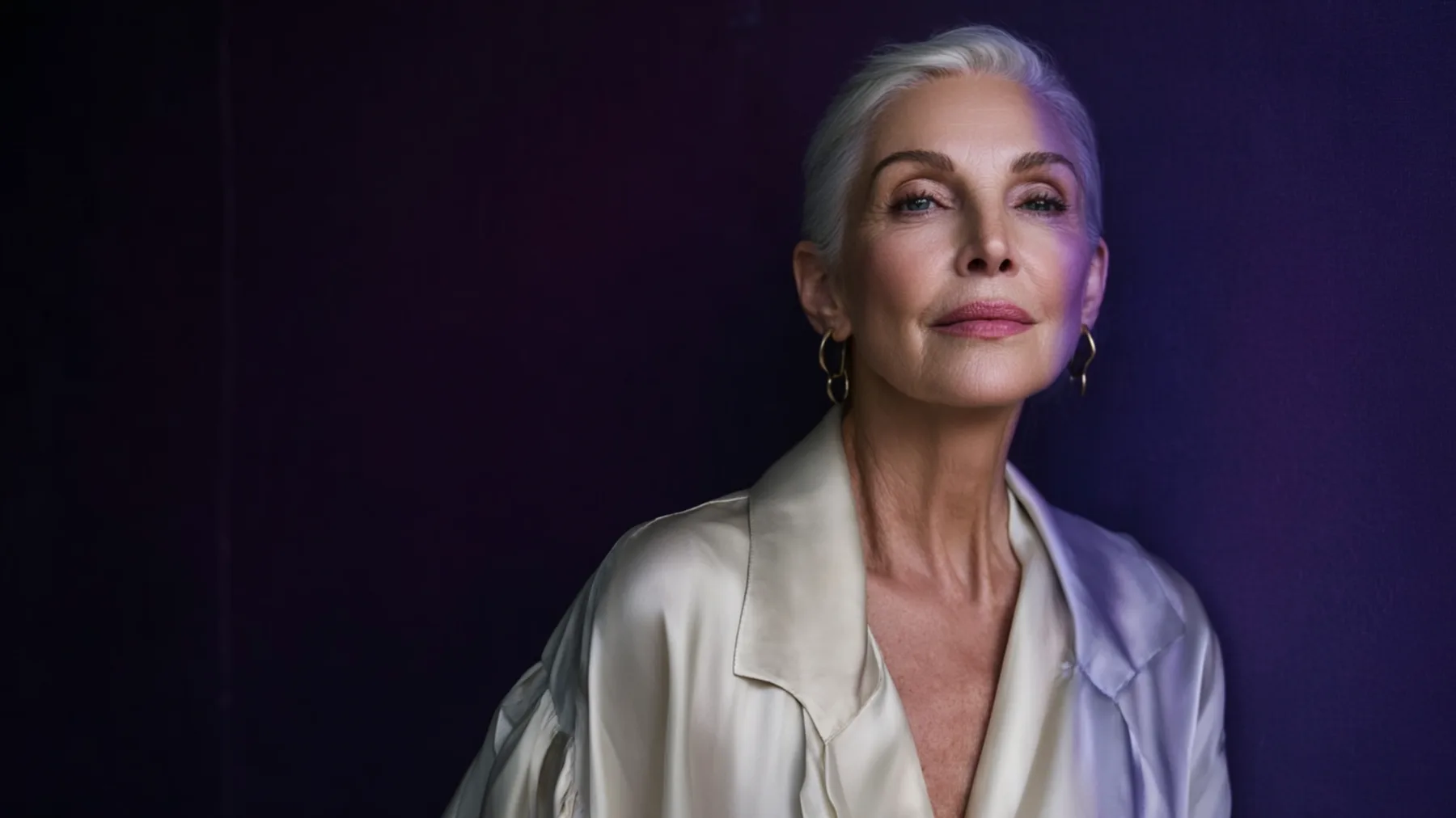 An elegant woman with silver hair wearing a light grey-silver satin blouse, looking into the distance against a dark purple backdrop