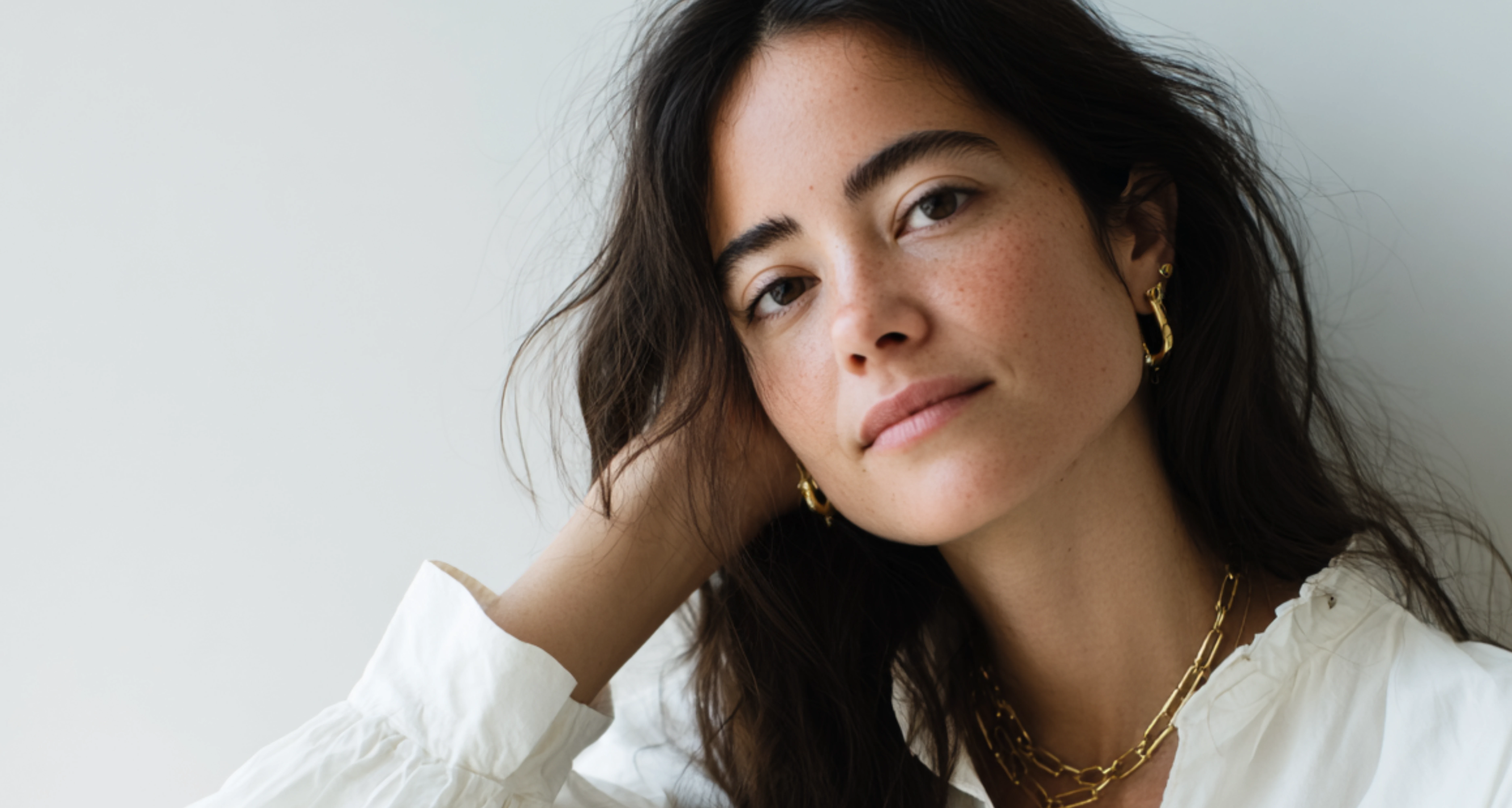 Person with long dark hair, gold jewelry, and a white blouse, looking calmly at the viewer
